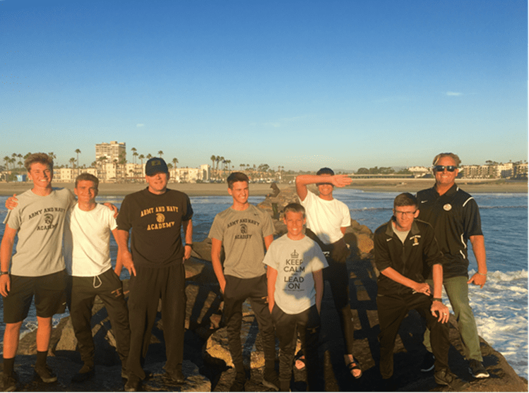 Army and Navy Academy surf team members and coaches pose on a coastal rock jetty with the ocean in the background at sunset.