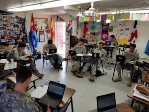 Cadets in camouflage uniforms work on laptops at desks in a JROTC classroom decorated with flags.