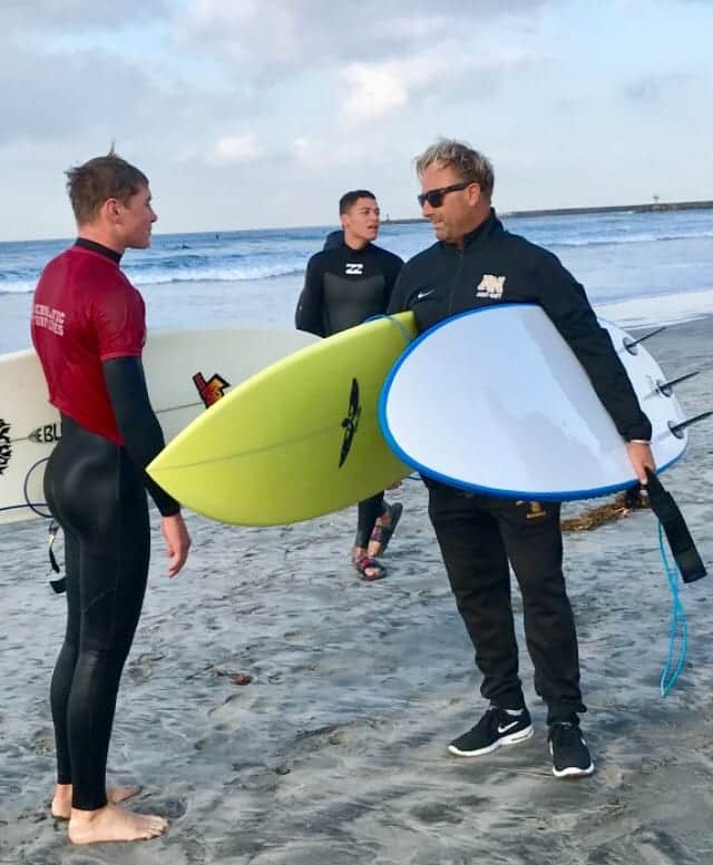 The Army and Navy Academy surf coach holds a surfboard while speaking with a cadet in a wetsuit on the beach before competing.