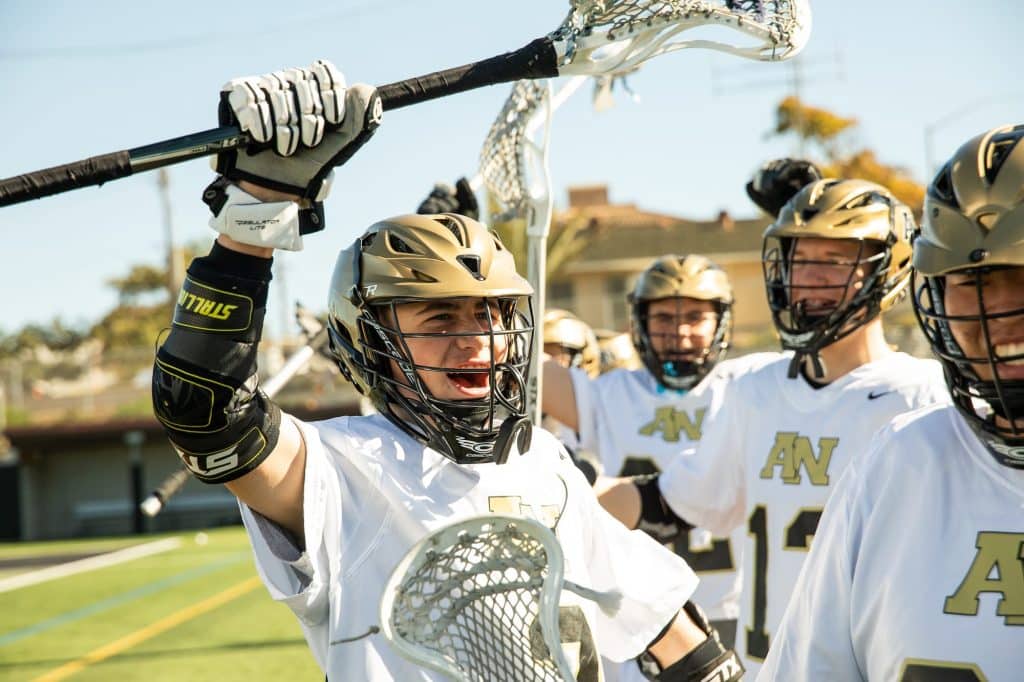 An Army and Navy Academy lacrosse player in white and gold uniform celebrates on the field with teammates during a game.