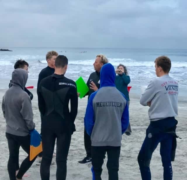 The Academy surf coach addresses a group of cadets on the beach before a Scholastic Surf Series competition on an overcast day.