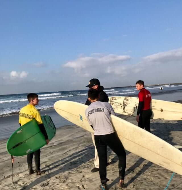 Cadet surfers in wetsuits carry their surfboards on the beach beside the ocean on a sunny morning.