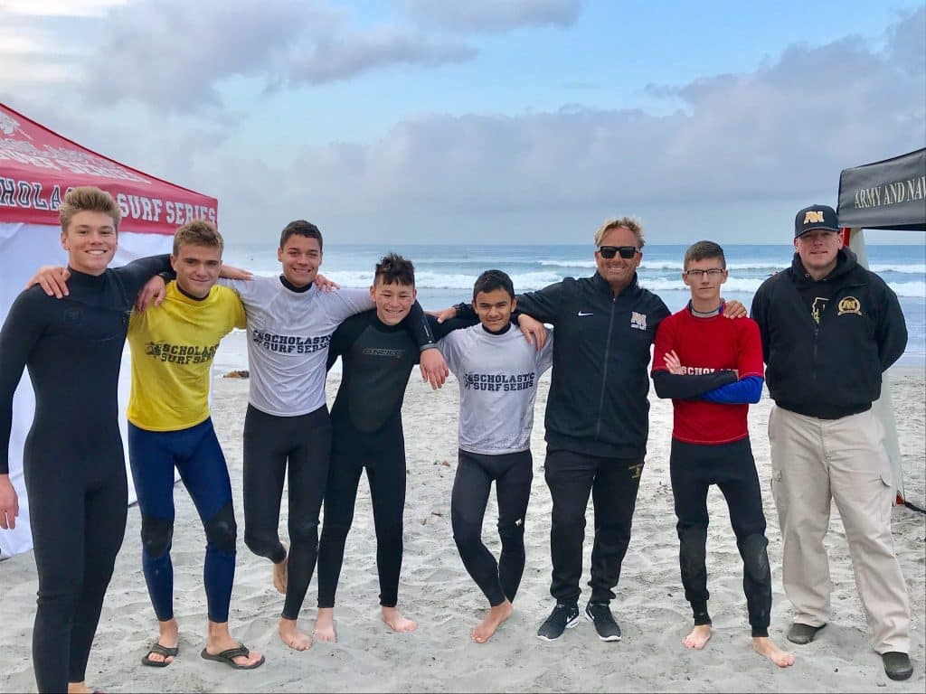 Army and Navy Academy surf team members and coaches pose on the beach at a Scholastic Surf Series competition, wearing wetsuits and team gear.
