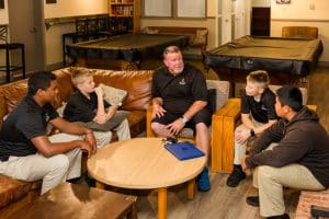 An adult mentor in a black polo leads a discussion with four cadets in a dormitory common room at Army and Navy Academy.
