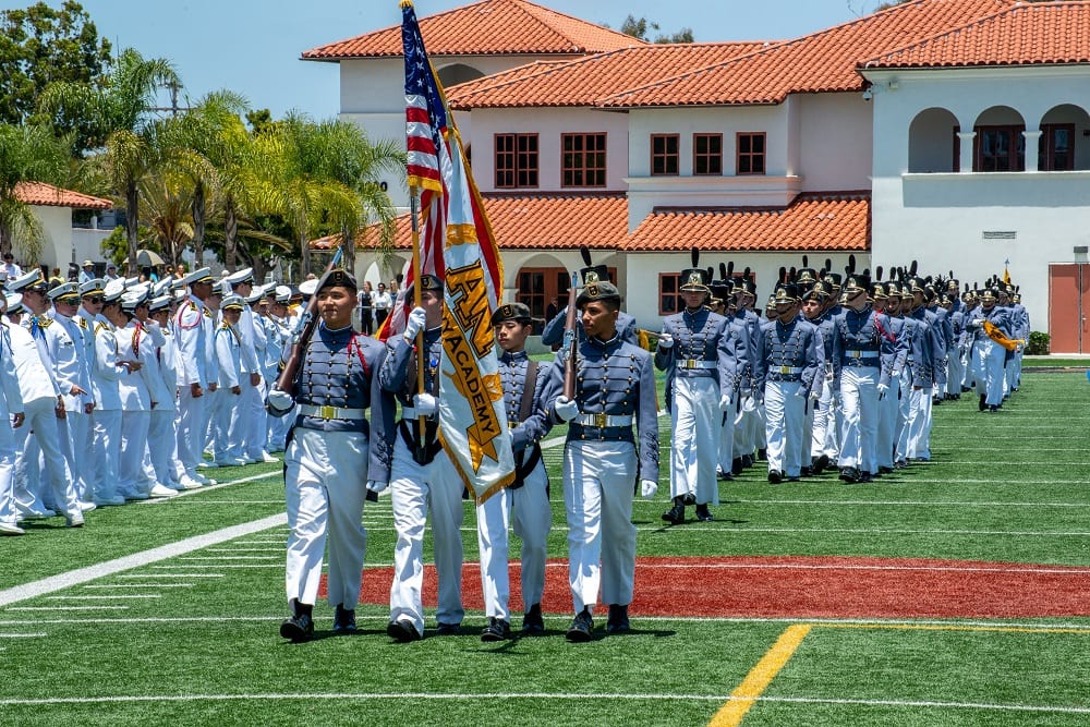 Army and Navy Academy cadets in dress white uniforms march in formation carrying the US flag and ANA banner during a parade ceremony.