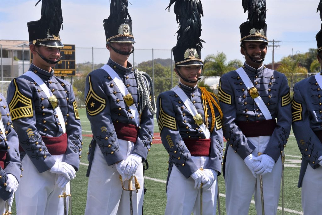 Cadets in full gray dress uniforms with feathered shakos stand in formation on the athletic field at Army and Navy Academy.