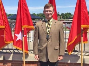 Major General Bartell wearing a Hall of Fame medal stands before red Army general officer flags at the ROTC Hall of Fame induction ceremony.