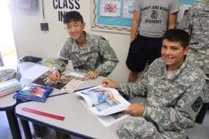 Two cadets in camouflage uniforms smile while reviewing a publication at a classroom desk.
