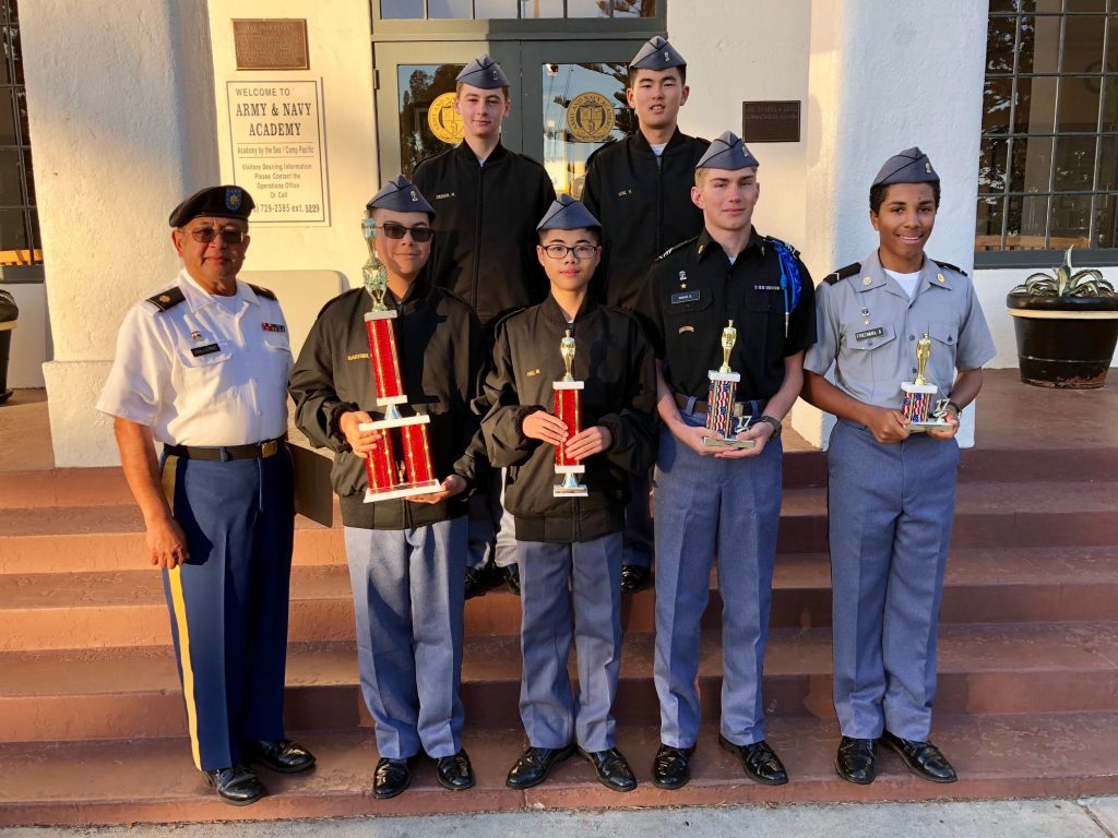 An instructor and seven cadets in JROTC uniforms pose on the Army and Navy Academy entrance steps holding drill competition trophies.