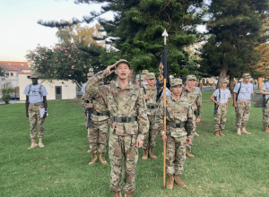 An Army and Navy Academy cadet in camouflage uniform salutes while leading a formation on the campus field during a JROTC drill.