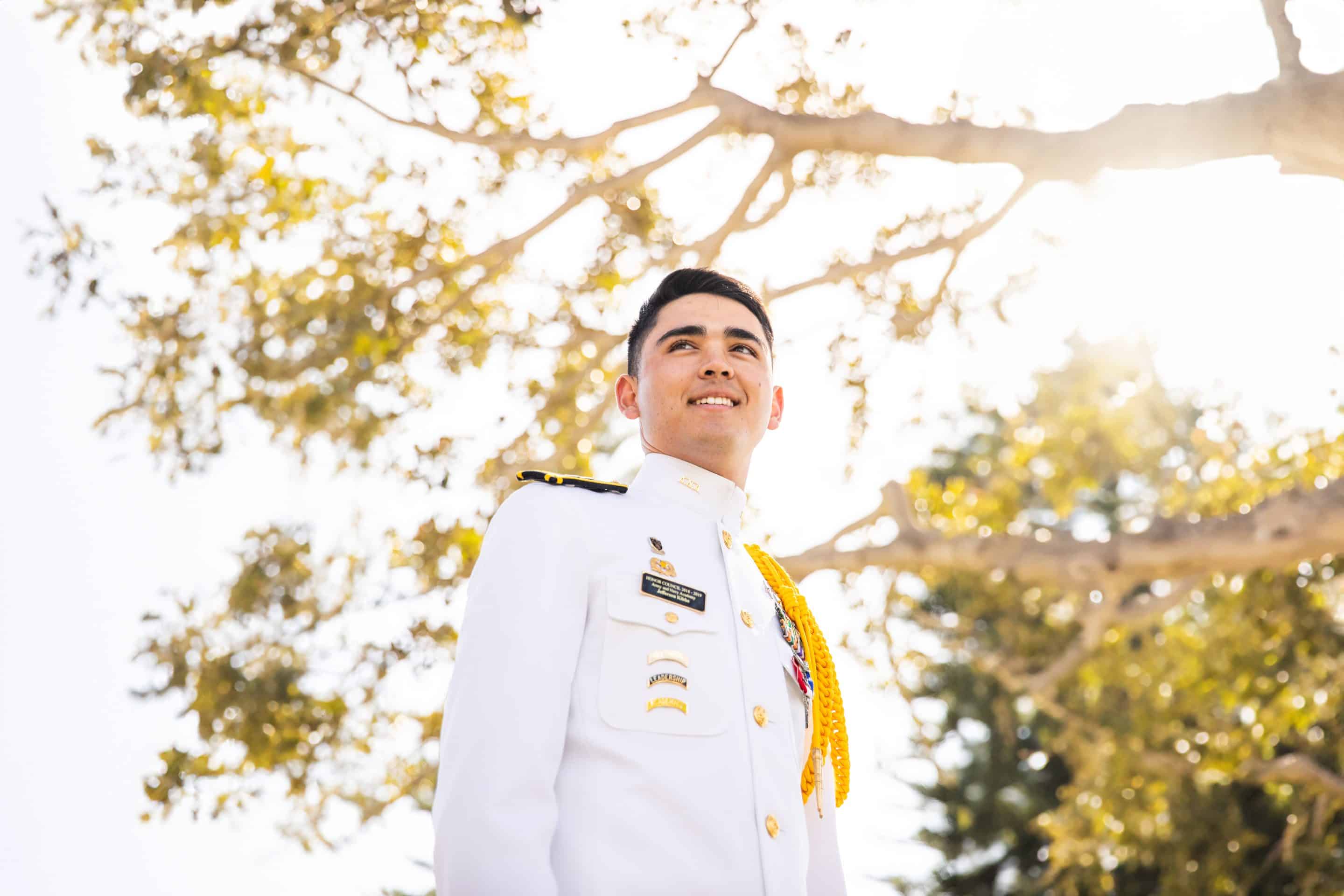 Army and Navy Academy cadet in white dress uniform with medals and gold aiguillette, smiling outdoors beneath sunlit trees.