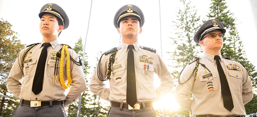 Three Army and Navy Academy cadets in dress gray uniforms with award ribbons stand at attention outdoors.