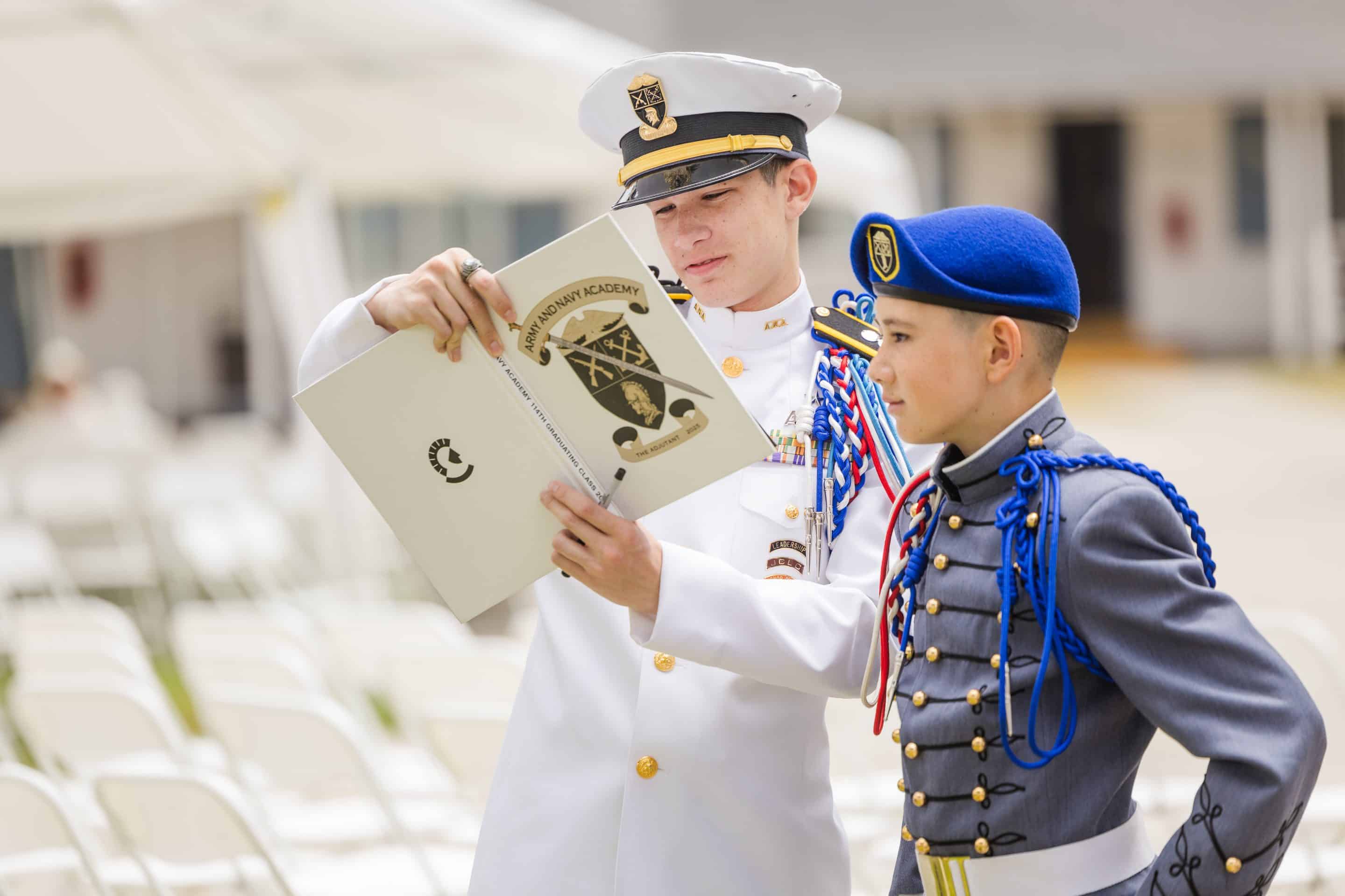 A senior cadet in white dress uniform and a younger cadet in full parade dress look together at the Army and Navy Academy yearbook.