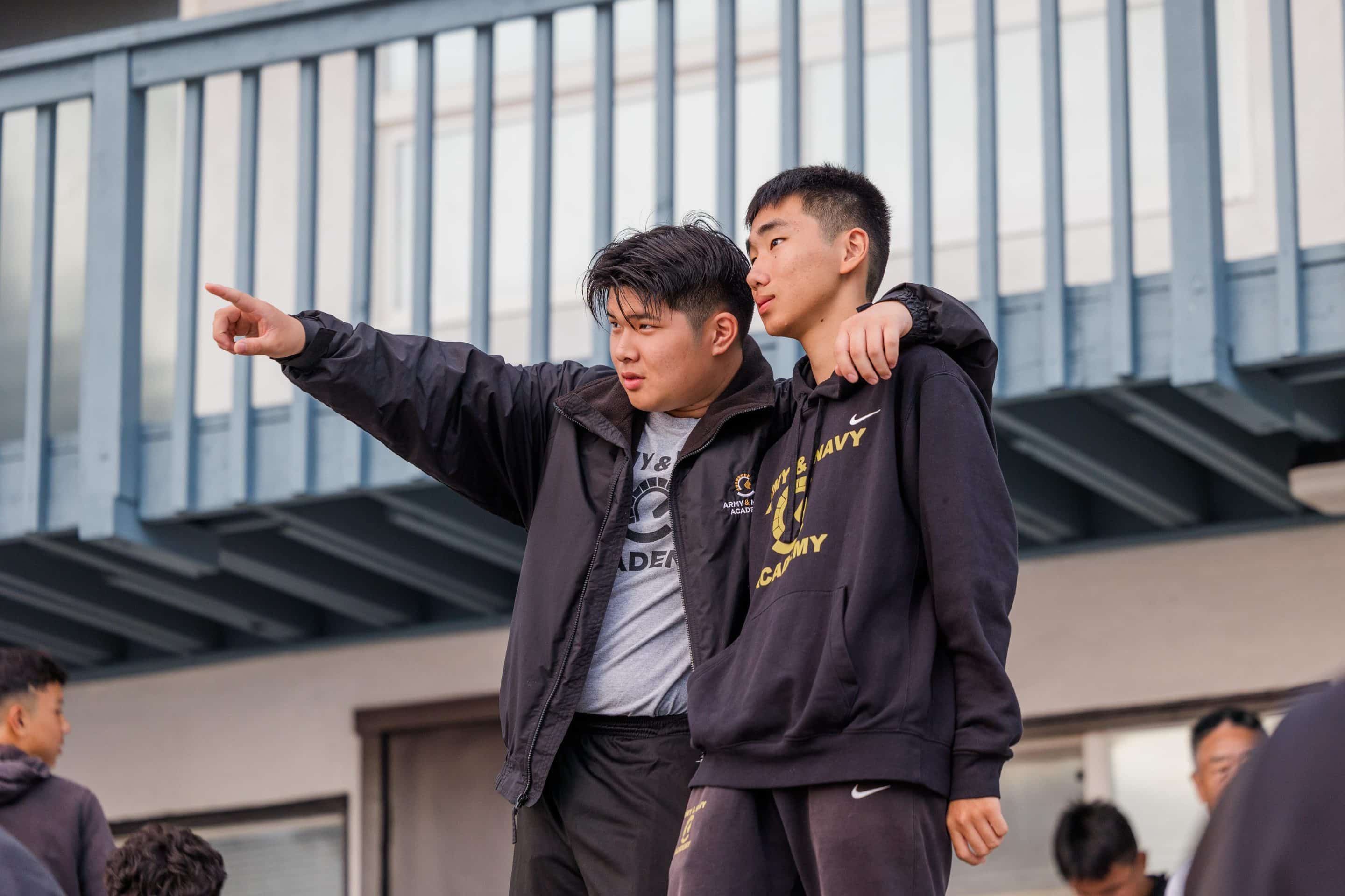 Two Army and Navy Academy cadets in casual gear stand together on campus, one pointing into the distance.