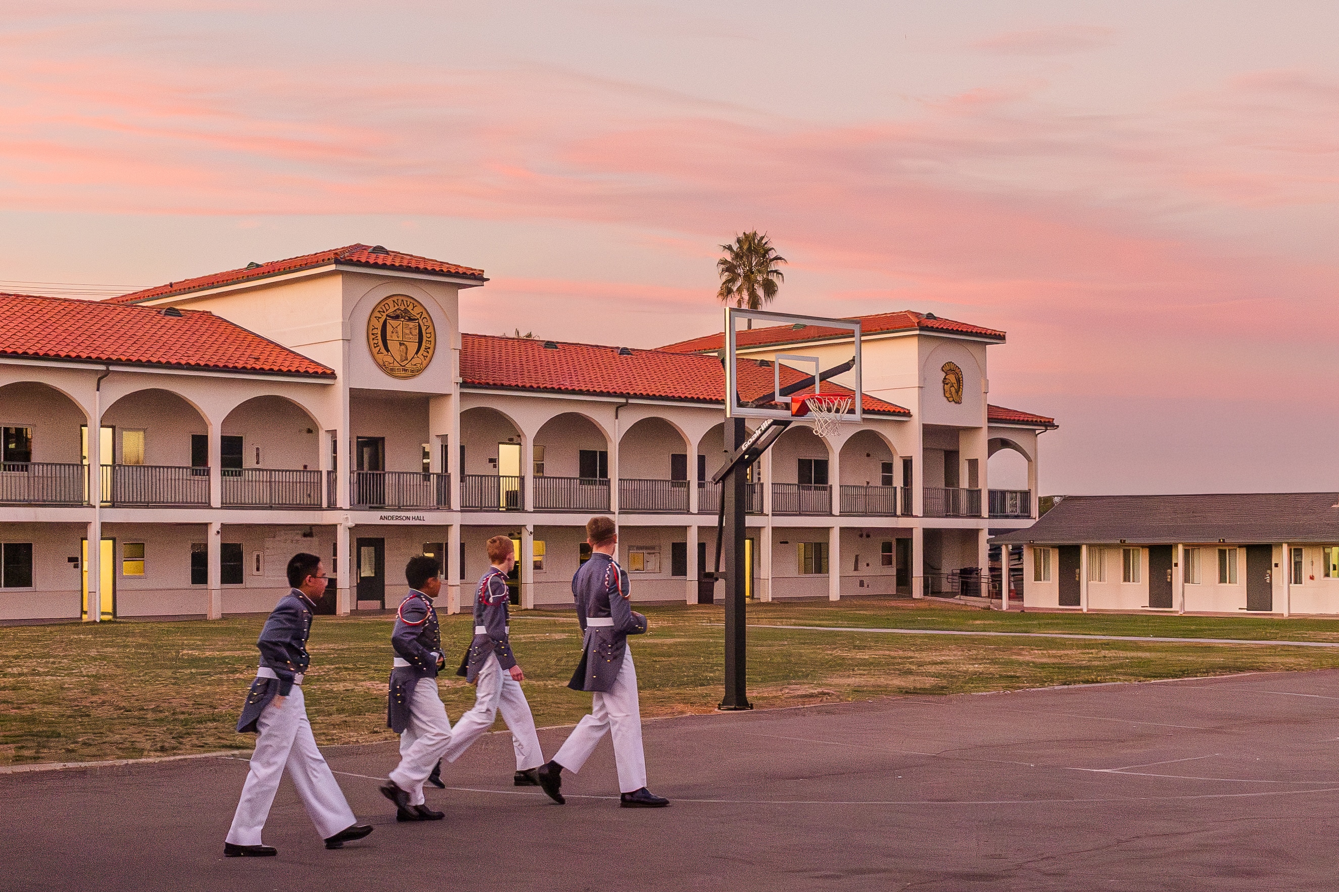 Army and Navy Academy cadets in dress white uniforms march across campus at sunset with a Spanish-style building behind them.