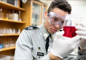 An Army and Navy Academy cadet in dress gray uniform examines a red liquid in a beaker during a science lab.