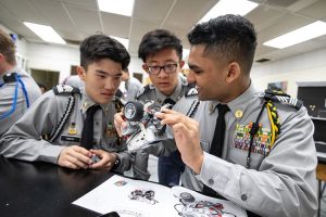Three cadets in gray uniforms collaborate over a small robot model during a STEM class at Army and Navy Academy.
