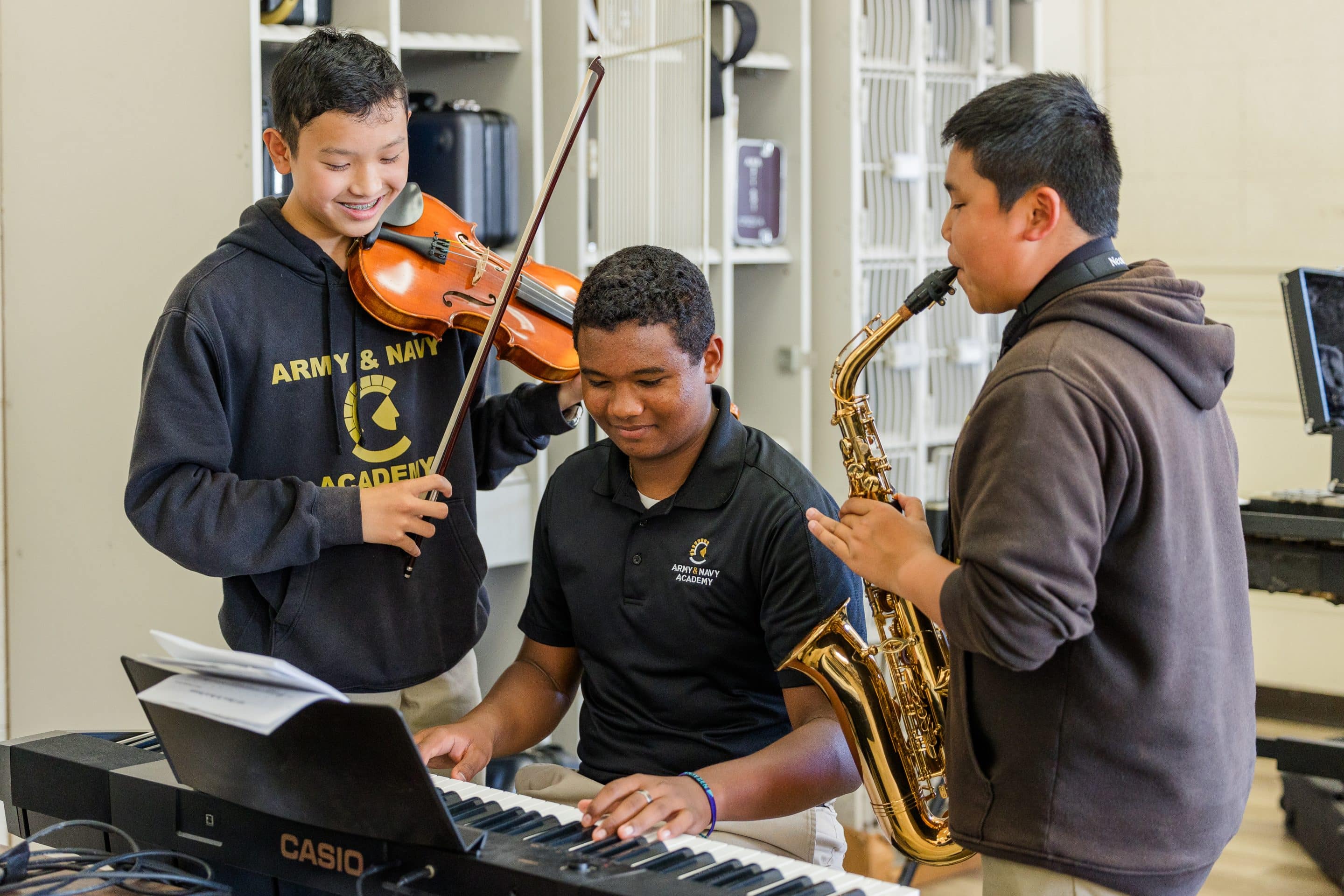 Three Army and Navy Academy cadets play violin, keyboard, and saxophone together in a campus music room.