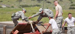 Cadets in camouflage uniforms assist each other over a wooden obstacle during a leadership challenge course at Army and Navy Academy.