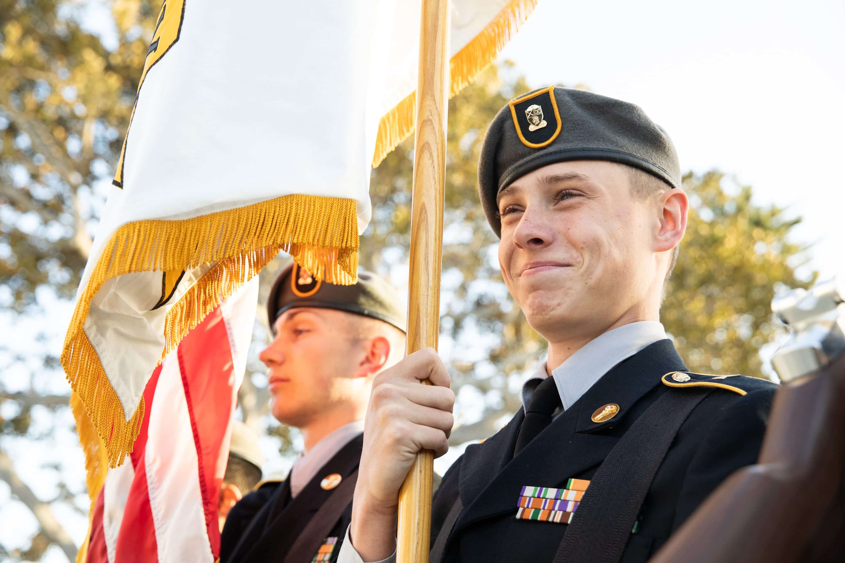 Army and Navy Academy cadet in Army dress uniform and green beret holds a ceremonial flag during an outdoor formation.