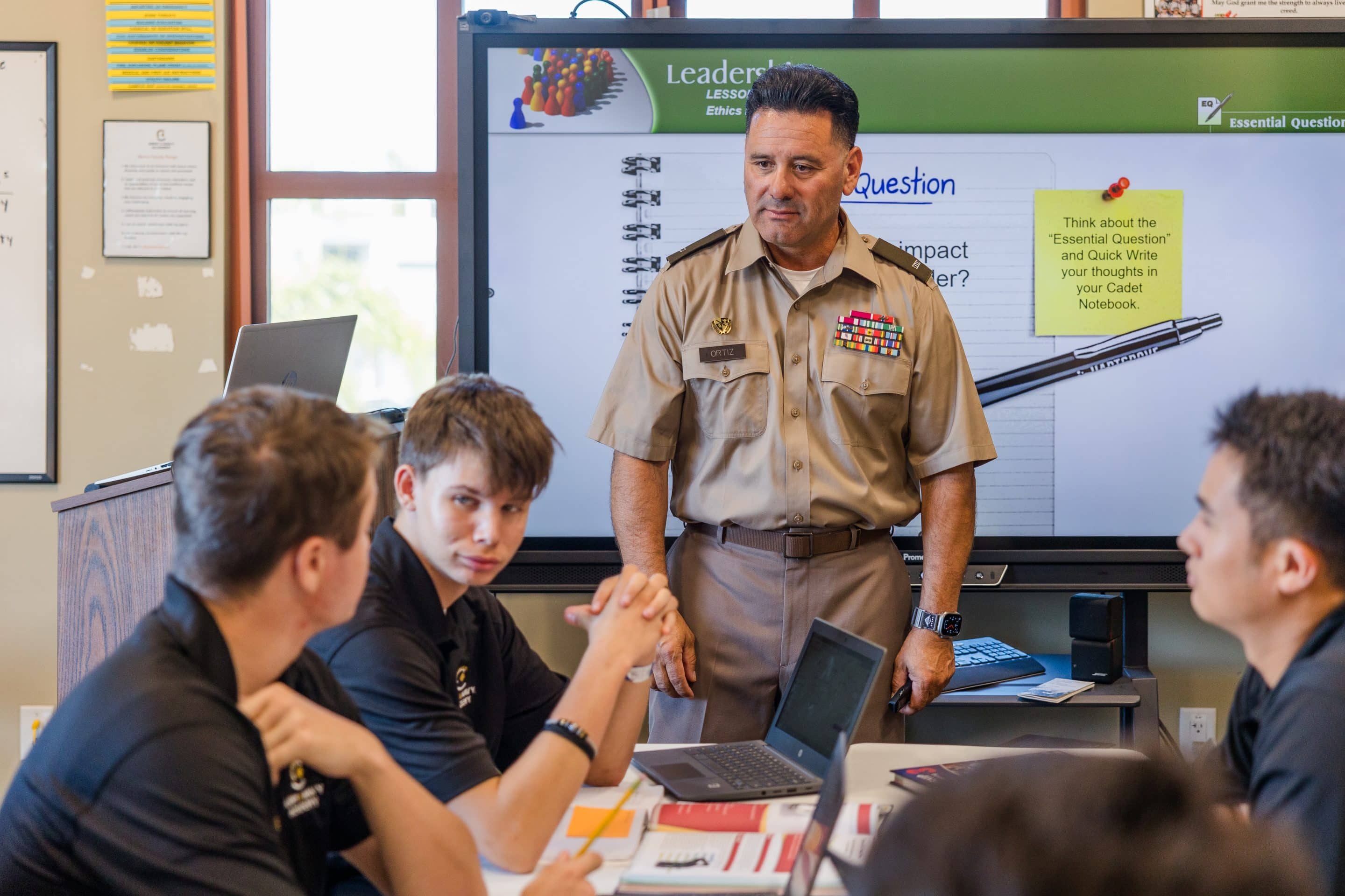A uniformed military instructor leads a leadership lesson at the smartboard for cadets seated at tables.