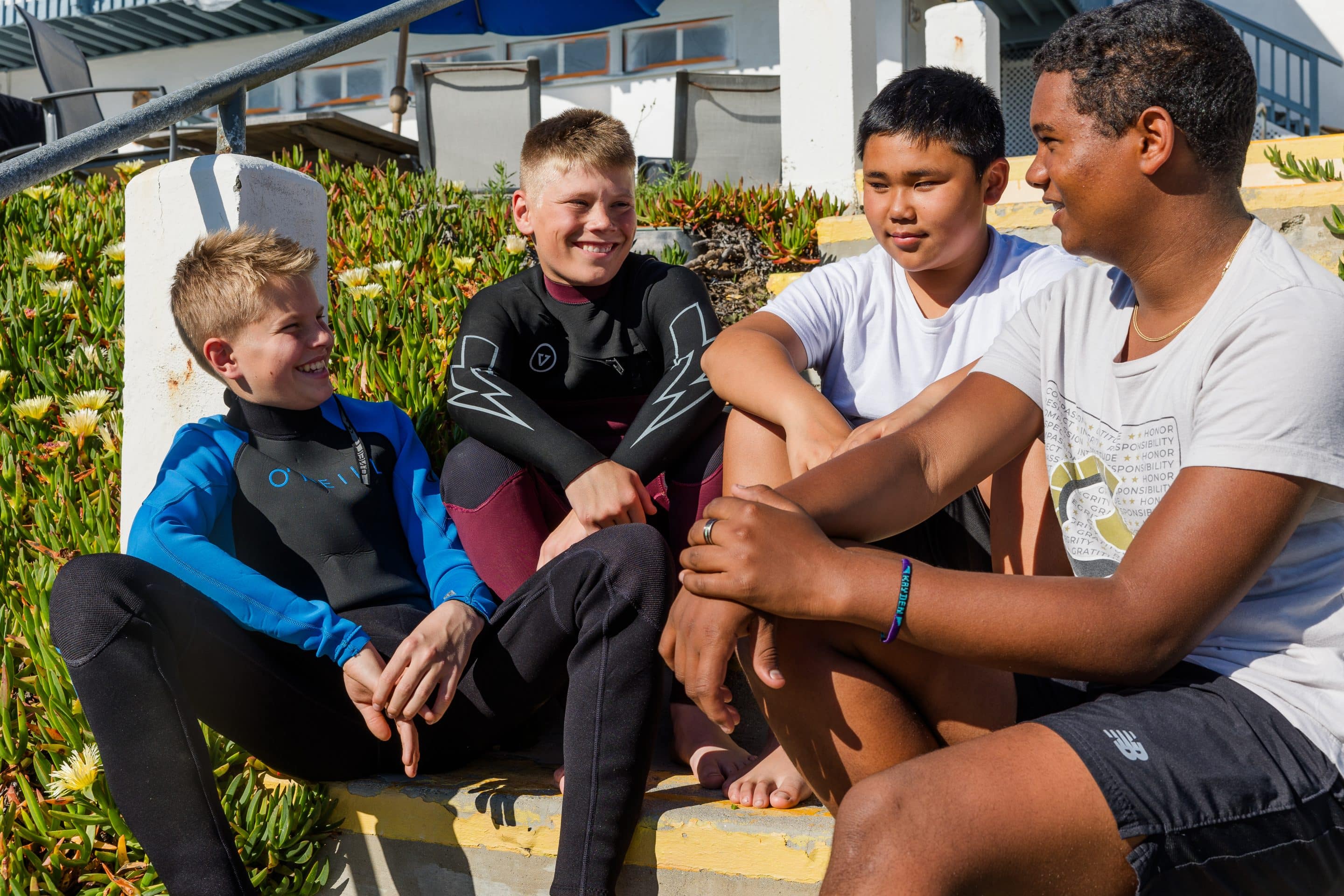 Four boys, two in wetsuits, sitting together near the beach at Army and Navy Academy during a shadow day visit.