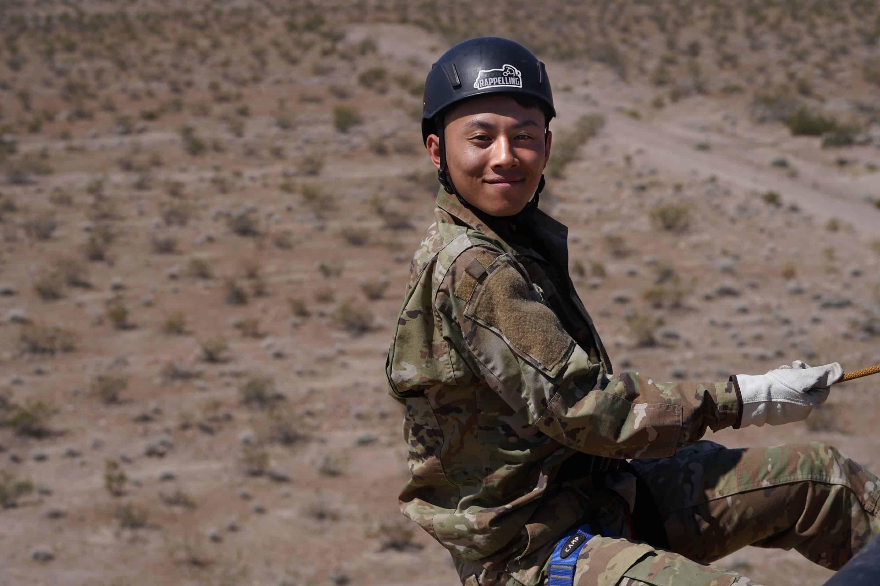 A cadet in camouflage uniform and rappelling helmet smiles while holding a rope during an outdoor adventure activity.