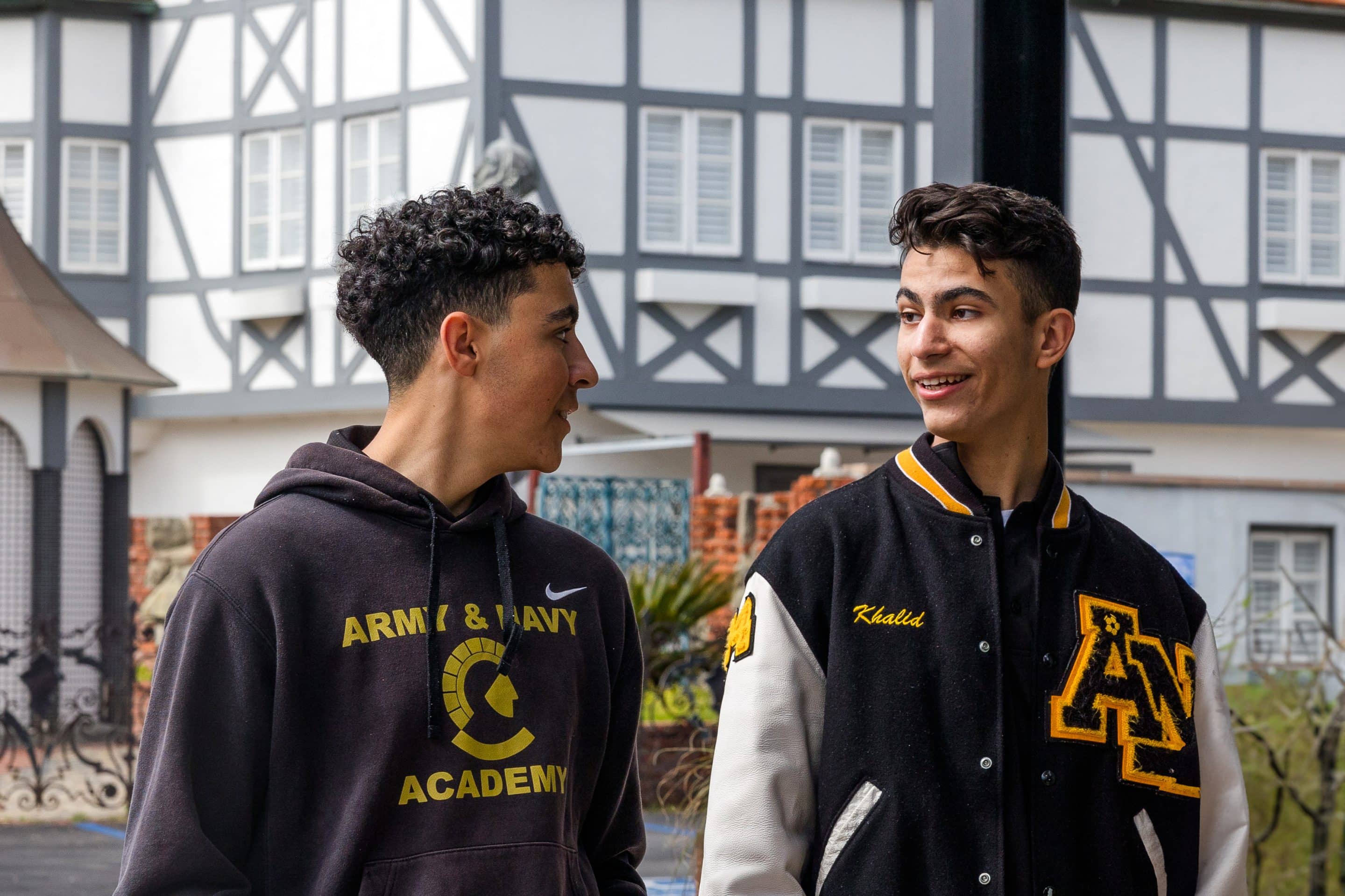 Two Army and Navy Academy cadets chat outside a Tudor-style campus building, one wearing an ANA varsity jacket.